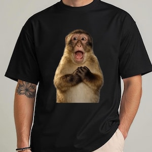 Puede incluir: Camiseta negra con un mono sorprendido, con la boca abierta y las manos juntas. El mono tiene pelaje marrón y beige. La camiseta es de color negro liso.