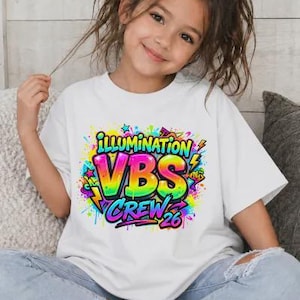 May include: White t-shirt with a colorful graphic that reads "ILLUMINATION VBS CREW 26". The design features bright colors, including yellow, green, blue, and pink, with a graffiti-style aesthetic. The shirt is worn by a young person.