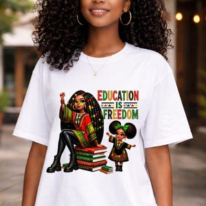 May include: White t-shirt featuring a colorful graphic with the text "EDUCATION IS FREEDOM." The graphic depicts two Black women, one seated on books labeled "Black History." The seated woman wears a red and green outfit, and the other wears a green bow.