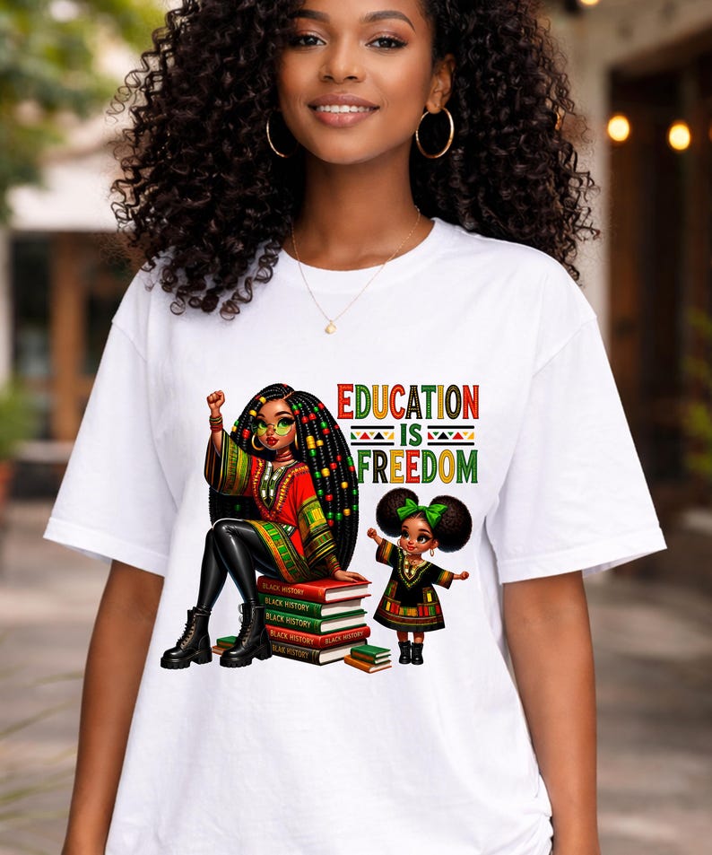 May include: White t-shirt featuring a colorful graphic with the text "EDUCATION IS FREEDOM." The graphic depicts two Black women, one seated on books labeled "Black History." The seated woman wears a red and green outfit, and the other wears a green bow.