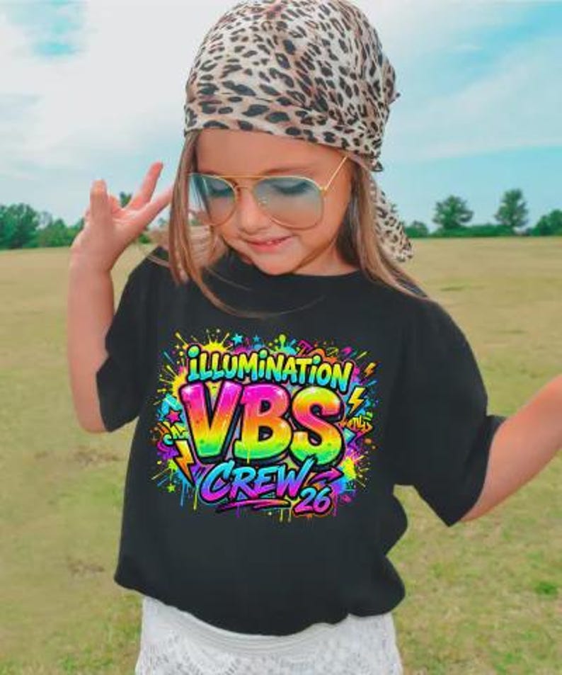 May include: A black t-shirt with the words "ILLUMINATION VBS CREW 26" in a colorful, graffiti-style design. The shirt is worn by a person wearing a leopard print headscarf and aviator sunglasses.