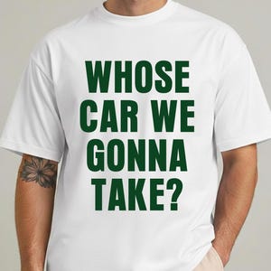 Può includere: T-shirt bianca con la scritta "WHOSE CAR WE GONNA TAKE?" in lettere maiuscole verde scuro. La maglietta ha un girocollo e maniche corte.