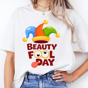 以下が含まれることがあります： 青、オレンジ、緑の要素を持つカラフルな道化師の帽子デザインが特徴の白いTシャツ。「BEAUTY FOOL DAY」のテキストは太字の赤色で、漫画の目と舌のグラフィックが描かれています。エイプリルフールに最適です。