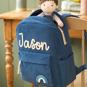 May include: A navy blue corduroy backpack featuring the name Jason embroidered in white script on the front pocket. A small fabric doll with brown hair and striped clothing sits inside the open main compartment. A small blue rainbow patch is attached.