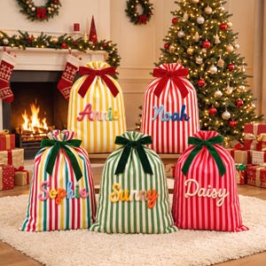 May include: Five fabric Christmas gift sacks with vertical stripes in yellow, red, green, and pink. Each sack features a velvet ribbon bow at the top and a personalised name in raised, colourful lettering: Annie, Noah, Sophie, Sunny, and Daisy.
