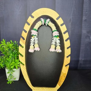 May include: A wooden display stand with an oval shape, featuring a black interior and a light brown frame. Two floral garlands with white, pink, and green accents are displayed. A small potted plant with green leaves is on the left.