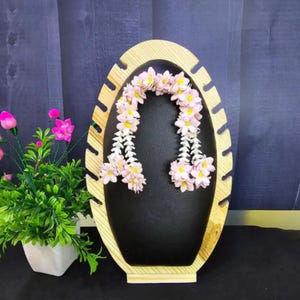 May include: A wooden jewelry display stand with an oval shape and a black interior. The stand features slots for hanging jewelry and is adorned with a garland of pink and yellow flowers. A small potted plant with pink flowers is on the left.