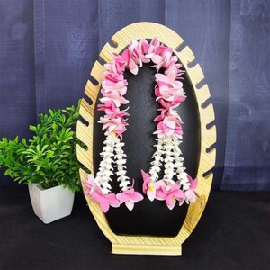 May include: A decorative wooden display stand holds two floral garlands. The garlands feature pink and white flowers, with a black background. The stand is made of light-colored wood and has an oval shape.