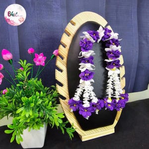 May include: A wooden display stand with a black backing holds two floral garlands. The garlands feature purple and white flowers. A potted plant with pink flowers and green leaves sits to the left of the stand. The background is a dark blue.