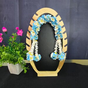May include: A wooden display stand with an oval-shaped opening, adorned with a garland of blue and white artificial flowers. The stand is made of light-colored wood and has a dark background. A small potted plant with pink flowers is on the left.