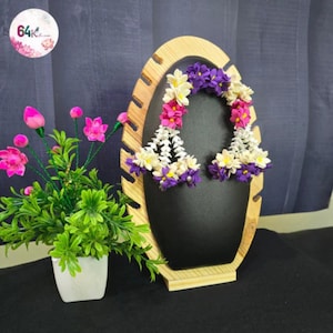 May include: A wooden earring display with a black center holds two floral earrings. The earrings feature white, pink, and purple flowers. A small potted plant with pink flowers and green leaves sits to the left.