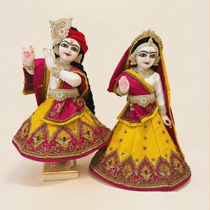 May include: Two ornate figurines dressed in vibrant yellow and magenta fabric garments with intricate gold embroidery. The figures feature detailed facial expressions, jewellery, and headpieces, standing in a pose with one hand raised in a gesture.