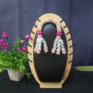 May include: A floral garland with white and pink flowers, displayed on a black stand with a wooden frame. The garland is adorned with pink and yellow accents. A small potted plant with pink flowers is in the background.