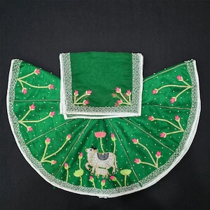 May include: A green embroidered outfit with a skirt and matching square scarf. The skirt is decorated with pink floral embroidery and a cow image. The edges are trimmed with white and gold accents.