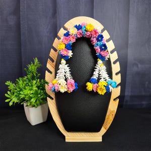 May include: A decorative floral garland featuring a vibrant mix of small blue, pink, yellow, and white flowers arranged in a semi-circular arch. Two dangling tassels made of white and multi-colored flowers hang from each end, accented by white beads.