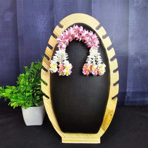 May include: A wooden jewelry display stand with a black oval center. The stand holds a pair of floral earrings with pink, white, and yellow accents. A small potted plant is to the left of the stand.