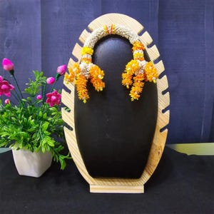 May include: A black jewelry display stand with a light wood frame. The stand is decorated with a garland of orange and white flowers. A small white pot with pink flowers and green leaves is on the left.