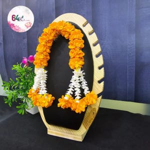 May include: A wooden display stand with a black interior holds a garland of orange and white flowers. The stand has a light wood frame with an oval shape and a decorative cutout design. A small plant is visible on the left.