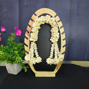 May include: A wooden jewelry display stand with an oval shape and cut-out design. A garland of white flowers with yellow centers hangs on the black background. A small potted plant with pink flowers sits to the left.