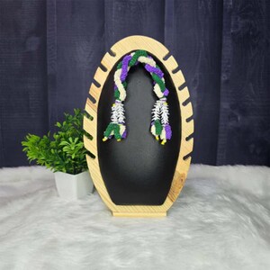 May include: A wooden jewelry display stand with an oval shape and a black interior. A colorful necklace with green, purple, and white accents hangs from the top. A small potted plant sits to the left of the stand.