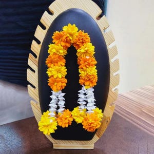 May include: A black oval display stand with a wooden frame, showcasing a garland of yellow and orange flowers with white accents. The floral arrangement is centered on the stand, creating a visually appealing display.