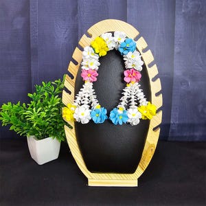 May include: A wooden jewelry display stand with a black surface, holding a colorful floral garland. The garland features white, yellow, pink, and blue flowers. A small potted plant is on the left.