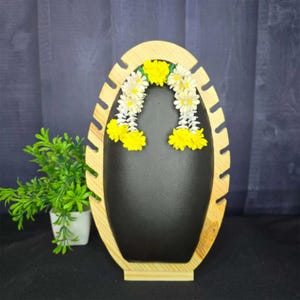 May include: Wooden jewelry display stand with a black backing and a light wood frame. The stand is decorated with a garland of white and yellow flowers. A small potted plant is visible on the left side of the image.