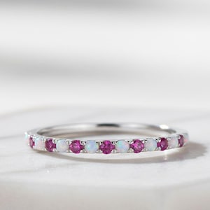 May include: A silver ring featuring alternating pink and white gemstones. The ring's design is set on a white surface, highlighting the gemstones' sparkle. A piece of jewellery, suitable for daily wear or special events. The gemstones are set in a band.