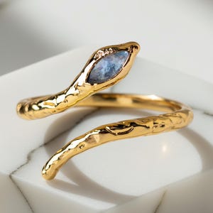May include: A gold-colored snake ring with a textured surface. The snake's head features a blue gemstone. The ring is coiled around a white marble block, creating a striking contrast. This unique piece is a statement accessory.