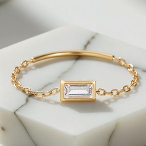 Peut inclure: Une bague en chaîne dorée avec une pierre précieuse rectangulaire et transparente sertie dans une lunette dorée. La bague est posée sur une surface géométrique blanche. La bague a une fine chaîne et une barre droite en haut.