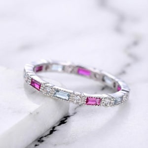 May include: A silver ring with alternating pink, light blue, and clear gemstones. The gemstones are rectangular and round, set in a continuous band. The ring is displayed on a white marble surface.