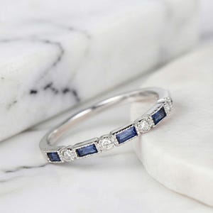 May include: A silver band ring with alternating rectangular blue and round clear gemstones. The ring's design is highlighted against a white marble backdrop, showcasing its elegant sparkle.