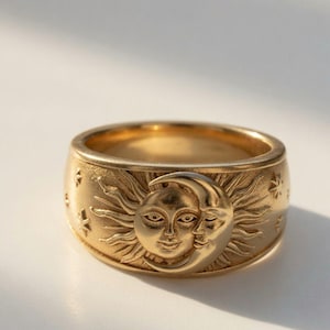 May include: A wide gold band ring featuring a raised relief design of a sun and a crescent moon face. The surface of the ring is textured with engraved stars and rays extending from the central celestial figures, finished in a matte gold tone.