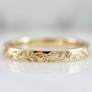 May include: A gold ring featuring a continuous engraved pattern of delicate leaves and small vines wrapping around the entire outer surface of the band. The metal has a polished, reflective finish that catches the light, highlighting the intricate details.