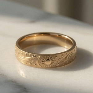 May include: A gold-toned metal ring featuring an engraved celestial pattern around the outer band. The design includes a central sun, a crescent moon, and various stars and shooting comets etched into the smooth, polished surface of the band.