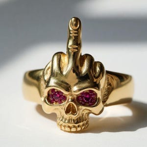 May include: A gold-colored ring featuring a skull design with ruby-colored eyes and a raised middle finger. The ring is a bold statement piece, showcasing intricate details and a unique, edgy aesthetic.