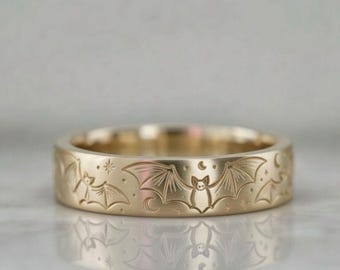 Puede incluir: Un anillo dorado con un patrón repetido de murciélagos, estrellas y lunas crecientes. El anillo tiene un acabado pulido y una anchura de banda de aproximadamente 0,64 cm.