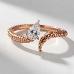 May include: A rose gold bypass ring featuring a textured, scale-like pattern along the band. One end of the open band terminates in a pear-shaped, faceted clear gemstone, while the other end tapers to a point, mimicking the appearance of a snake.