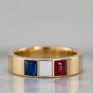 May include: A gold ring with a flat band, featuring three square insets of blue, white, and red. The ring's design is reminiscent of a flag, with a polished, reflective surface.