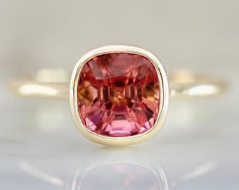 May include: A close-up of a gold ring featuring a large, cushion-cut, pink gemstone. The gemstone is set in a bezel setting, and the band is a simple, polished gold. The ring is displayed on a reflective surface.