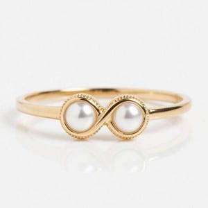 May include: A gold-colored ring featuring two white pearls set in an infinity symbol design. The ring has a delicate band and a textured border around each pearl, creating an elegant and timeless piece of jewelry.