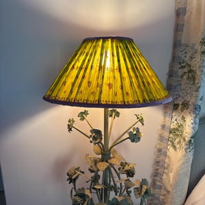 May include: A decorative table lamp with a green and yellow patterned lampshade. The lamp has a floral design with a metal base and a purple trim on the lampshade. The lamp is on a blue surface.