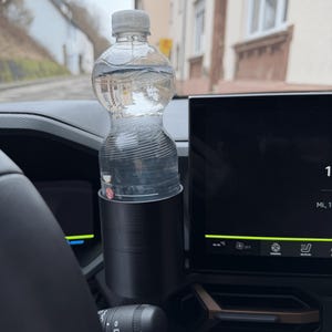 Könnte beinhalten: Eine transparente Plastikwasserflasche mit grauem Deckel steht in einem schwarzen Getränkehalter in einem Auto. Das Armaturenbrett und das Lenkrad des Autos sind sichtbar, mit einem verschwommenen Hintergrund von Gebäuden und einer Straße.