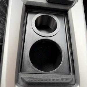 Puede incluir: Inserto de consola de coche negro, impreso en 3D, con dos portavasos circulares de diferentes tamaños. El inserto encaja en una consola de coche gris, con un soporte para teléfono visible arriba. Diseño funcional y moderno.