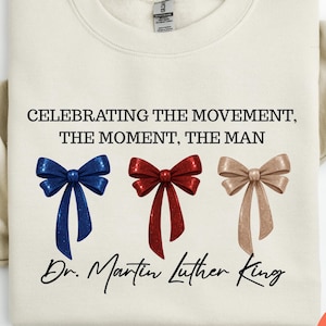 Peut inclure: Sweat-shirt cr&egrave;me avec le texte "CELEBRATING THE MOVEMENT, THE MOMENT, THE MAN" et "Dr. Martin Luther King." En dessous, trois n&oelig;uds bleus, rouges et beiges. Le sweat-shirt a un col rond.