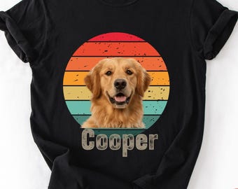 May include: Black t-shirt with a golden retriever dog design. The dog is set against a retro sunset design with red, orange, yellow, and teal stripes. The name "Cooper" is printed below the dog in a vintage font. The shirt is made of a soft material.