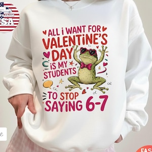 May include: White sweatshirt with a Valentine's Day graphic. The design features a frog wearing heart-shaped sunglasses and a bow tie, with text that reads "All I want for Valentine's Day is my students to stop saying 6-7."