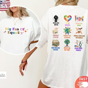 Op de afbeelding: Wit T-shirt met de tekst "Big Fan Of Equality" in kleurrijke letters. De achterkant van het shirt heeft verschillende afbeeldingen en zinnen, waaronder "Black Lives Matter", "Love is Universal" en "Empower All Women".