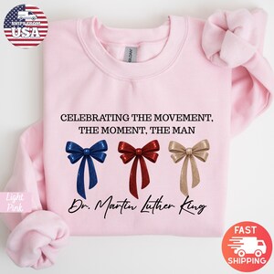 Peut inclure: Sweat-shirt rose clair avec le texte "CELEBRATING THE MOVEMENT, THE MOMENT, THE MAN" et "Dr. Martin Luther King." Comprend trois graphiques de n&oelig;uds papillon bleus, rouges et dor&eacute;s. Le sweat-shirt a des manches longues.