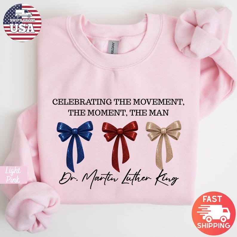 Peut inclure: Sweat-shirt rose clair avec le texte "CELEBRATING THE MOVEMENT, THE MOMENT, THE MAN" et "Dr. Martin Luther King." Comprend trois graphiques de n&oelig;uds papillon bleus, rouges et dor&eacute;s. Le sweat-shirt a des manches longues.